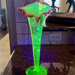 Puede incluir: Un jarrón de vidrio verde vibrante con una parte superior acampanada y una base redonda. El jarrón presenta delicados diseños florales y un borde teñido de rojo. El jarrón es un artículo decorativo, probablemente para exhibir flores.