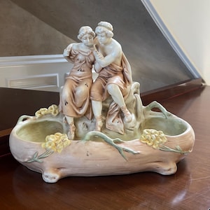 May include: A porcelain planter featuring a sculpted scene of two figures seated on a rock. The planter is decorated with floral details and has a light brown and green glaze.