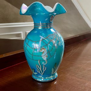 May include: A blue glass vase with a white rim and a seahorse design. The vase has a silver glitter finish and is decorated with a seahorse, seaweed, and a net.