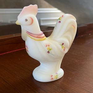 May include: A white milk glass rooster figurine with hand-painted pink, yellow, and green floral details. The rooster has a pink comb and wattle and a black eye.