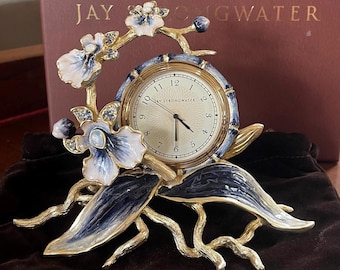 Może przedstawiać: Ozdobny zegar o tematyce kwiatowej, z kremową tarczą i złotymi wskazówkami. Zegar otoczony jest złotymi gałązkami i niebiesko-białymi kwiatami emaliowanymi. Na tarczy zegara widnieje napis "JAY STRONGWATER".