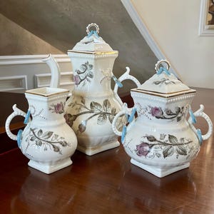 Puede incluir: Un juego de té de porcelana blanca con un diseño floral. El juego incluye una tetera, una jarra de crema y un azucarero. Las piezas están decoradas con delicadas rosas rosadas y hojas verdes. Las asas están acentuadas con cintas azules.