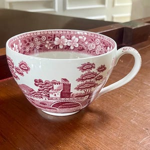 Puede incluir: Una taza de té de cerámica blanca con un patrón de transferencia de color rosa oscuro. El exterior muestra una escena paisajística con un puente de piedra y edificios, mientras que el borde interior está decorado con un borde floral y pequeños motivos circulares.