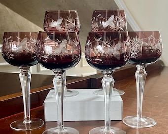 May include: Six wine glasses with a deep burgundy bowl and clear stem. Each glass features an etched design of a bird in flight and other decorative elements. The glasses are arranged on a wooden surface, suggesting a formal setting.