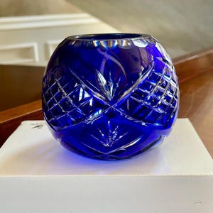 Puede incluir: Un jarrón redondo de cristal azul cobalto con intrincados patrones tallados de diamantes y estrellas. El jarrón se encuentra sobre una superficie blanca, mostrando sus cualidades brillantes y reflectantes. Un elemento decorativo.