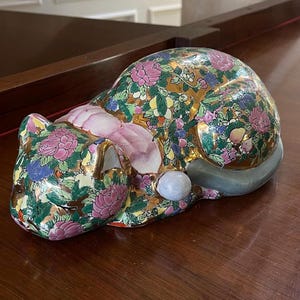 May include: A porcelain cat figurine with a gold background and colorful floral pattern. The cat is lying down with its head resting on its paws.