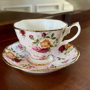 May include: A porcelain teacup and saucer set featuring a pale pink background with a delicate white lace-like pattern. The set is decorated with vibrant yellow and deep red rose blossoms and green leaves, finished with a polished gold trim.