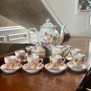 May include: A complete vintage tea set featuring a floral design. The set includes a teapot, sugar bowl, creamer, and several teacups with saucers. The porcelain pieces have gold trim and a pink and white floral pattern. The set is arranged on a dark wooden surface.