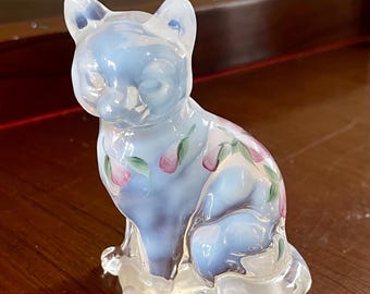 May include: A light blue glass cat figurine with pink floral accents. The cat is in a seated position with its eyes closed. The figurine is made of translucent glass, allowing light to pass through. A decorative item.