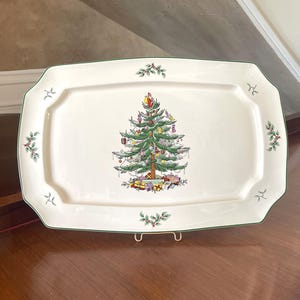 Puede incluir: Una bandeja de servir rectangular de cerámica color crema con un borde verde oscuro. El centro presenta un árbol de Navidad decorado con adornos coloridos y regalos envueltos en la base. Pequeñas ramas de acebo decoran el borde.