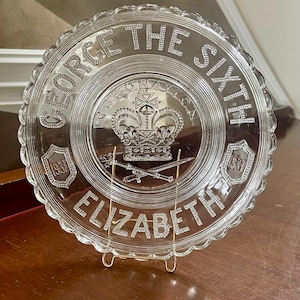 Op de afbeelding: Een herdenkingsbord van helder glas met de in reliëf aangebrachte tekst GEORGE THE SIXTH en ELIZABETH. Het midden toont een kroon, het woord CORONATION en gekruiste zwaarden. Twee schilden aan de zijkanten bevatten het jaartal 1937.