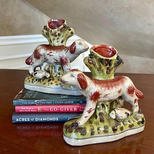 May include: Two ceramic dog figurines, each featuring a white and reddish-brown spotted dog standing on a green and yellow mottled base next to a tree stump-shaped candle holder. The figurines are placed on a stack of three books.