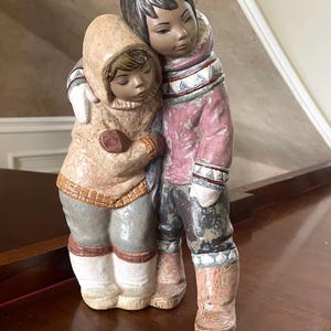 Puede incluir: Una figura de cerámica que representa a dos figuras con ropa de invierno. Una figura abraza a la otra. Las figuras llevan chaquetas con capucha, manoplas y botas. La paleta de colores incluye tonos de rosa, marrón, gris y blanco.