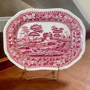 May include: An oval ceramic platter with a scalloped edge, featuring a deep red transferware pattern of a landscape with a stone bridge, trees, and buildings, set against a white background, displayed on a gold-toned wire stand.