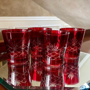 Pode incluir: Quatro copos de cristal vermelho rubi com desenhos geométricos e florais gravados. Os copos são exibidos em uma superfície espelhada, refletindo seus padrões intrincados. Uma adição clássica a qualquer coleção de bar.