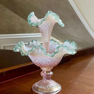 May include: A two-tiered glass vase with a pink and iridescent finish. The vase has a scalloped rim and a diamond pattern. The top tier is a single trumpet-shaped vase. The vase is sitting on a wooden surface.