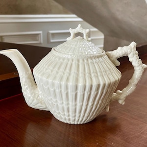 May include: A white ceramic teapot with a lid and a decorative handle. The teapot has a textured, ribbed design and a curved spout. The handle and lid finial have a branch-like appearance. The teapot is sitting on a wooden surface.