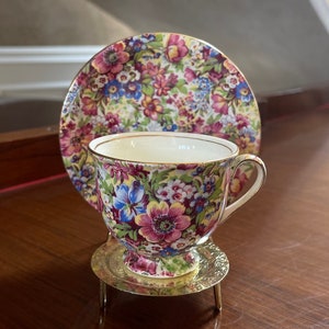Puede incluir: Un juego de taza y platillo de té floral con ribete dorado. La taza y el platillo están decorados con un patrón floral colorido. La taza está sobre un soporte de metal dorado con tres patas.