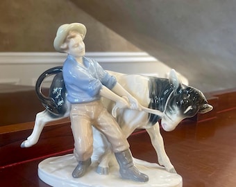 May include: A porcelain figurine of a young person in a straw hat and blue shirt leading a black and white calf by a rope.