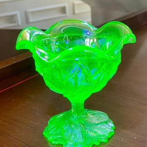 Puede incluir: Una copa de cristal de color verde neón vibrante con un borde ondulado y una base de pedestal. La superficie exterior está decorada con un patrón de hojas en relieve que se extiende desde la base hasta el cuenco, creando un aspecto texturizado.