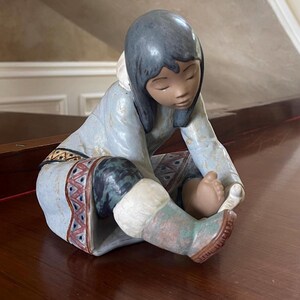 Può includere: Statuina in ceramica di una persona in abbigliamento Inuit tradizionale, con parka e stivali blu. La figura è seduta, guardando in basso i suoi piedi. Gli stivali presentano un motivo decorativo rosso, bianco e blu. Oggetto decorativo.