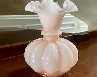 Op de afbeelding: Een lichtroze glazen vaas met een gerimpelde bovenkant en een bolvormig, getextureerd lichaam. De vaas staat op een houten oppervlak.