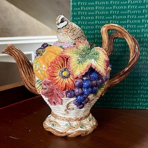 Puede incluir: Tetera de cerámica decorativa con un pájaro en la parte superior. La tetera está adornada con un diseño floral y frutal, incluyendo uvas, una calabaza y una flor grande. El asa y el pico tienen una textura similar a la madera.