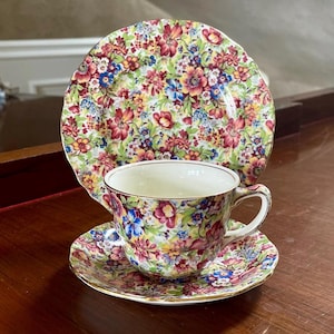 Könnte beinhalten: Ein florales Teetassen- und Untertassen-Set mit einem passenden floralen Teller. Das Set hat einen weißen Hintergrund mit einem bunten Blumenmuster in den Farben Rosa, Rot, Gelb, Blau und Grün.