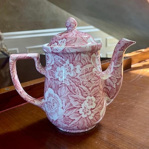 Puede incluir: Tetera de cerámica con tapa, asa y boquilla. Presenta un estampado floral de flores y hojas blancas sobre fondo rojo. La tetera está colocada sobre una superficie de madera.