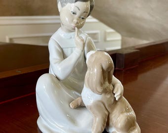 May include: A porcelain figurine of a girl with a finger to her lips, sitting with a brown and white dog. The girl is wearing a white dress and has brown hair. The dog is sitting on its hind legs and has its head tilted to the side.