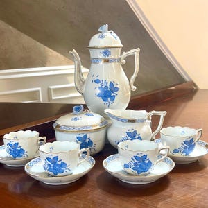 May include: A white porcelain tea set with blue floral designs and gold trim. The set includes a coffee pot, sugar bowl, creamer, and six teacups with saucers. The tea set is displayed on a wooden surface.