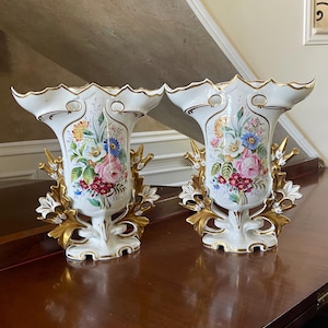 Może przedstawiać: Para białych porcelanowych wazonów z falistymi brzegami i złotymi wykończeniami. Każdy wazon przedstawia kolorowy bukiet kwiatów z przodu i jest ozdobiony dekoracyjnymi, złoconymi motywami liści i gałązek wychodzącymi od podstawy na boki.