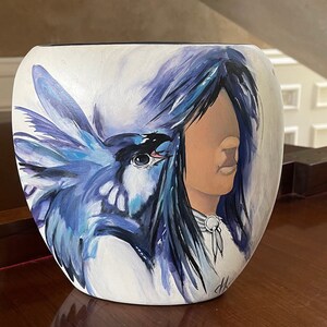 May include: White ceramic vase with a painted design featuring a Native American woman and a blue bird. The woman's face is in profile, with long dark hair. The bird is painted in shades of blue and black.