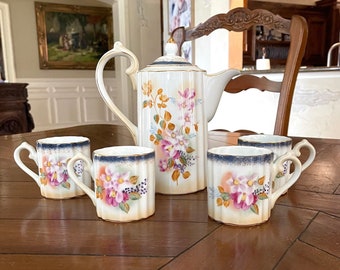 May include: A white porcelain teapot and four matching teacups with floral designs in shades of pink, yellow, and blue. The teapot has a gold trim and a blue band around the top. The teacups have a gold trim and a blue band around the bottom.