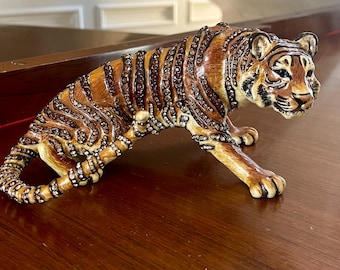 May include: A detailed figurine of a tiger in a crouching pose. The tiger is primarily brown with black stripes and is adorned with small, sparkling embellishments. The figurine is placed on a polished wooden surface.
