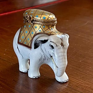 Könnte beinhalten: Eine weiße Elefantenfigur aus Keramik mit dekorativem Deckel. Der Deckel und der Rücken des Elefanten sind mit goldenen und blauen Akzenten verziert. Der Elefant hat detaillierte Merkmale, einschließlich Stoßzähnen und bemalten Augen.