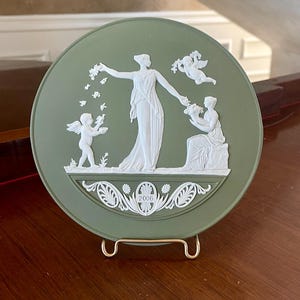Könnte beinhalten: Eine runde, salbeigrüne dekorative Platte mit einem weißen Reliefdesign. Das Design zeigt eine zentrale weibliche Figur, Cherubinen und florale Elemente. Das Jahr "2006" ist unten eingraviert. Die Platte wird auf einem goldfarbenen Metallständer präsentiert.