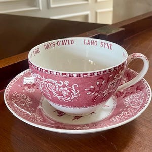 Puede incluir: Un juego de taza y plato de cerámica con un patrón floral en rosa oscuro y blanco. El borde interior de la taza muestra el texto 'FOR DAYS O'AVLD LANG SYNE' en letras de color rojo oscuro. El juego incluye un asa a juego y un borde decorativo.