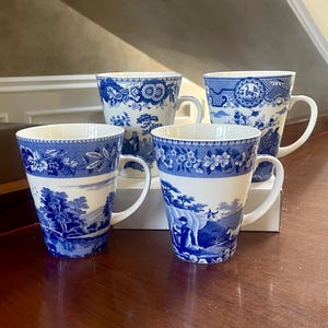May include: Four ceramic mugs featuring a white base with blue transferware patterns. Each mug displays detailed pastoral scenes, including trees, figures, and animals, with decorative floral borders around the rims and handles.