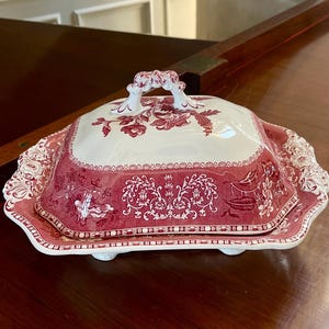 以下が含まれることがあります： 赤と白のセラミック製の蓋付きサービングディッシュ。この皿は花柄と風景模様が特徴です。蓋には取っ手があり、皿にはスカラップエッジがあります。この皿は木製の表面にあります。