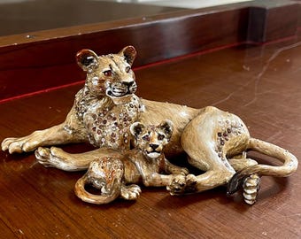 May include: A decorative figurine of a lioness and two cubs. The lioness and cubs are lying down, with the lioness in the center and the cubs on either side. The figurine is a light brown color with jewel accents.