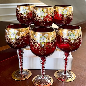 May include: Six red wine glasses with gold rims and floral designs. The bowls are a deep red, with gold accents and white floral patterns. The stems are clear with red and white twisted detailing, and the bases are gold.