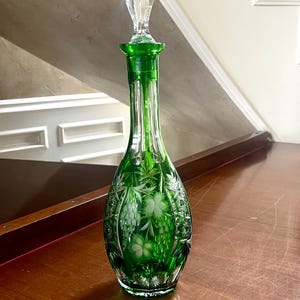 Puede incluir: Una jarra de vidrio verde con un tapón de vidrio transparente. La jarra tiene un cuello largo y un cuerpo bulboso con diseños grabados de flores y uvas. El tapón es facetado. La jarra está sobre una superficie de madera.
