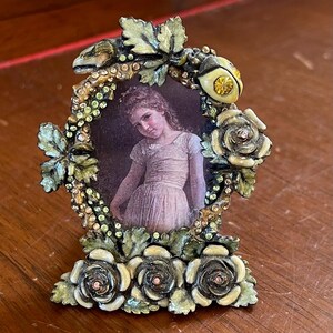 Puede incluir: Un marco de fotos ovalado de metal con un borde decorativo de hojas verdes, rosas de color crema y pequeños diamantes de imitación de color ámbar. El marco contiene el retrato de una persona joven con un vestido claro. La base está adornada con tres rosas.