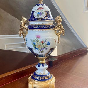 Könnte beinhalten: Eine weiße Porzellanurne mit Deckel, verziert mit tiefblauen und goldenen Zierbändern, Blumenmotiven und zwei goldfarbenen Puttenhenkeln. Das Stück steht auf einem goldfarbenen Sockel und ist mit einem detaillierten goldenen Knauf versehen.