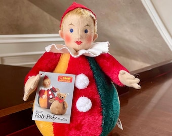 Könnte beinhalten: Eine Steiff Roly-Poly Replica Puppe. Die Puppe trägt ein farbenfrohes Outfit mit roten, grünen und gelben Abschnitten. Sie hat einen roten Hut, einen weißen Rüschenkragen und ein Etikett mit der Aufschrift "Steiff Roly-Poly Replica".