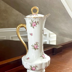 Może przedstawiać: Biały dzbanek z porcelany z złotym uchwytem i pokrywką. Dzbanek jest ozdobiony różowymi różami i zielonymi liśćmi.