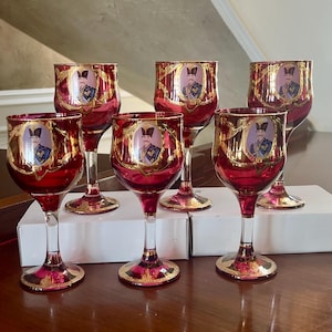 Puede incluir: Juego de siete copas de vino tinto con detalles dorados. Cada copa presenta un retrato enmarcado en oro. Las copas tienen un cuenco rojo, un tallo transparente y una base dorada. Se exhiben sobre una superficie de madera.