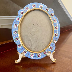 May include: An oval porcelain picture frame with a blue and gold background. The frame is decorated with pink roses and gold accents. The frame has three legs and is standing on a wooden surface.