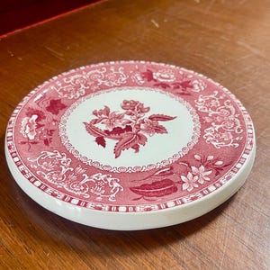 May include: A round ceramic coaster featuring a white center with a pink floral design, surrounded by a wide pink border decorated with intricate floral patterns and a repeating geometric motif along the outer edge of the circular piece.